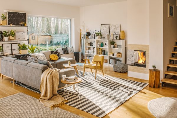 Blanket thrown on grey corner sofa in white living room interior with fresh plants, carpet, fireplace and simple posters